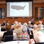 A woman speaks into a microphone in front of a screen while 20 people sit and listen